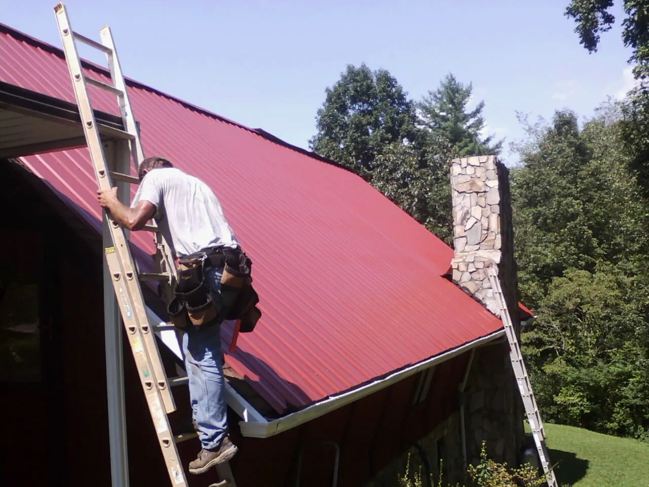 Roofer on a ladder beside a red metal roof and stone chimney — Soffit & Fascia Repair by Roofing Brothers in Scandia